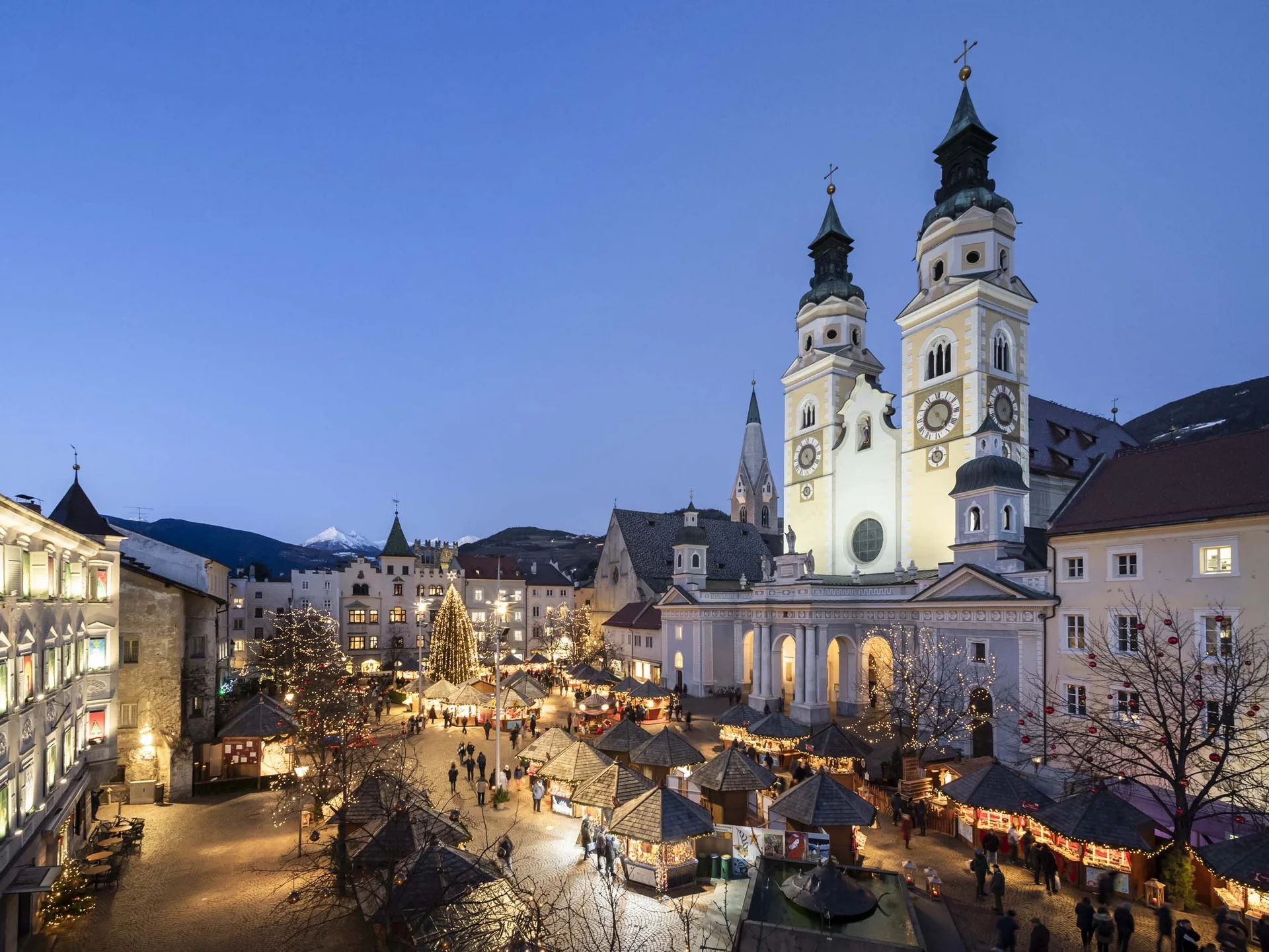 Der Lodenwirt in Vintl | Hotel, Residence & Restaurant Beleuchteter Weihnachtsmarkt auf einem Platz vor einer Kirche mit zwei Türmen in der Dämmerung