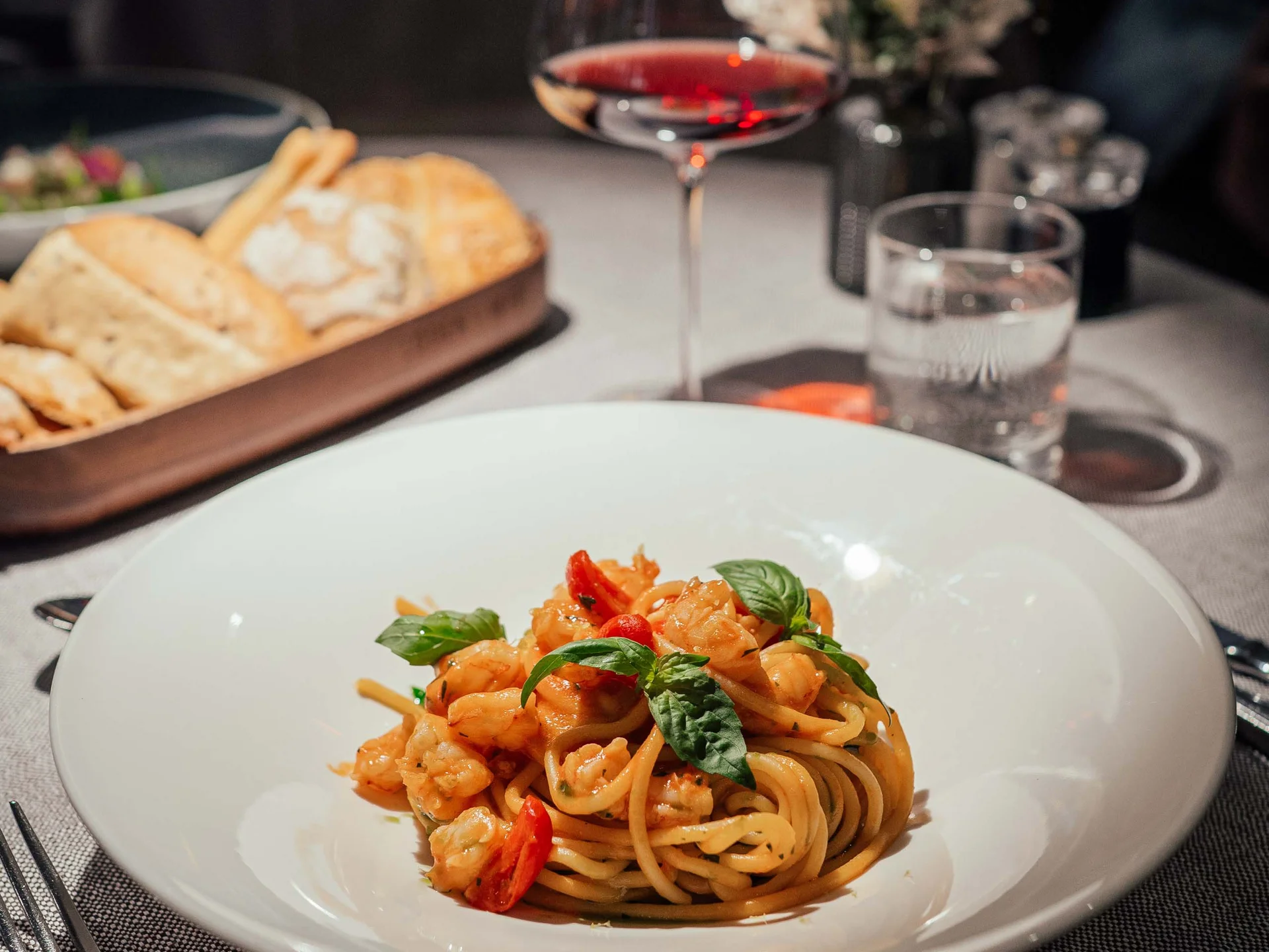 Der Lodenwirt in Vintl | Hotel, Residence & Restaurant Teller mit Pasta und Garnelen, Glas Rotwein und Brotkorb auf gedecktem Tisch