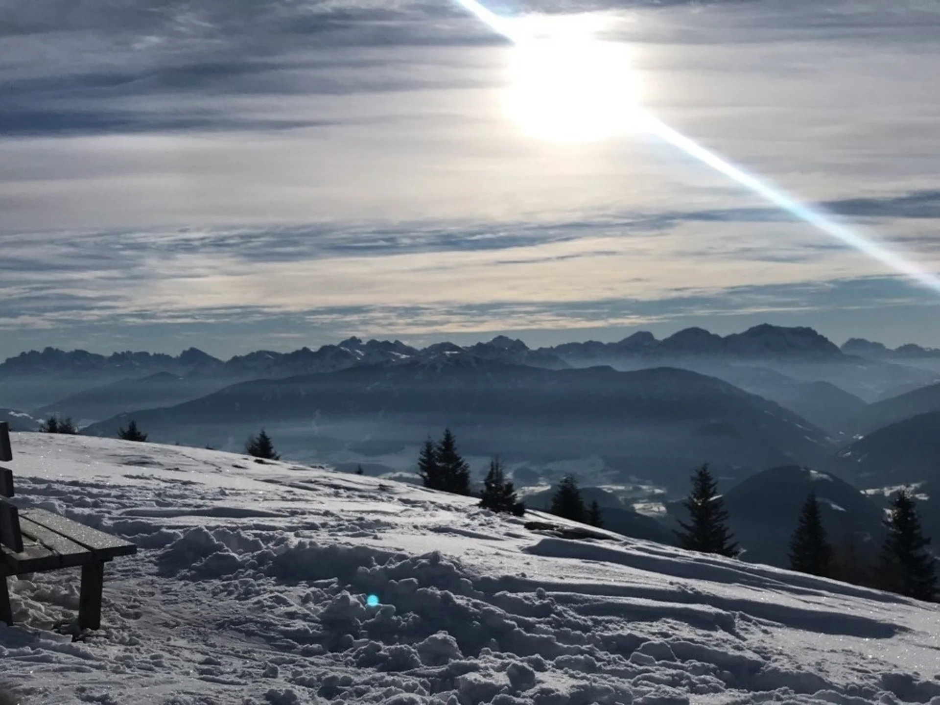 Der Lodenwirt in Vintl | Hotel, Residence & Restaurant Holzbank auf einem verschneiten Berghang mit Blick auf ein Gebirge im Sonnenlicht