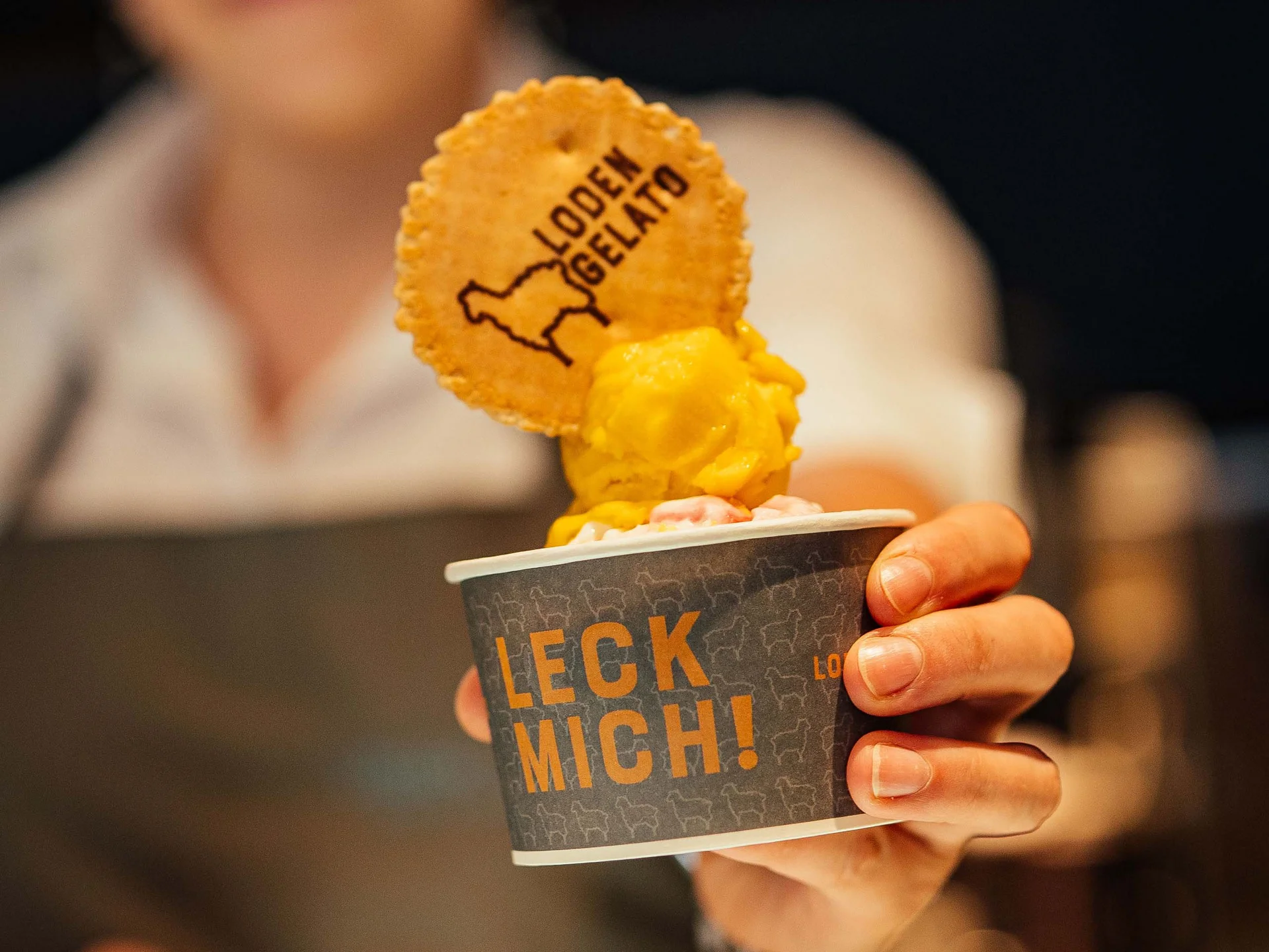 Der Lodenwirt in Vintl | Hotel, Residence & Restaurant Person hält Becher mit gelbem Eis und Keks mit Loden Gelato Schrift