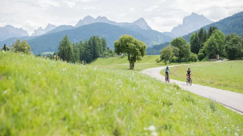 Der Lodenwirt in Vintl | Hotel, Residence & Restaurant Zwei Radfahrer auf einem Weg durch grüne Wiesen mit Bergen im Hintergrund