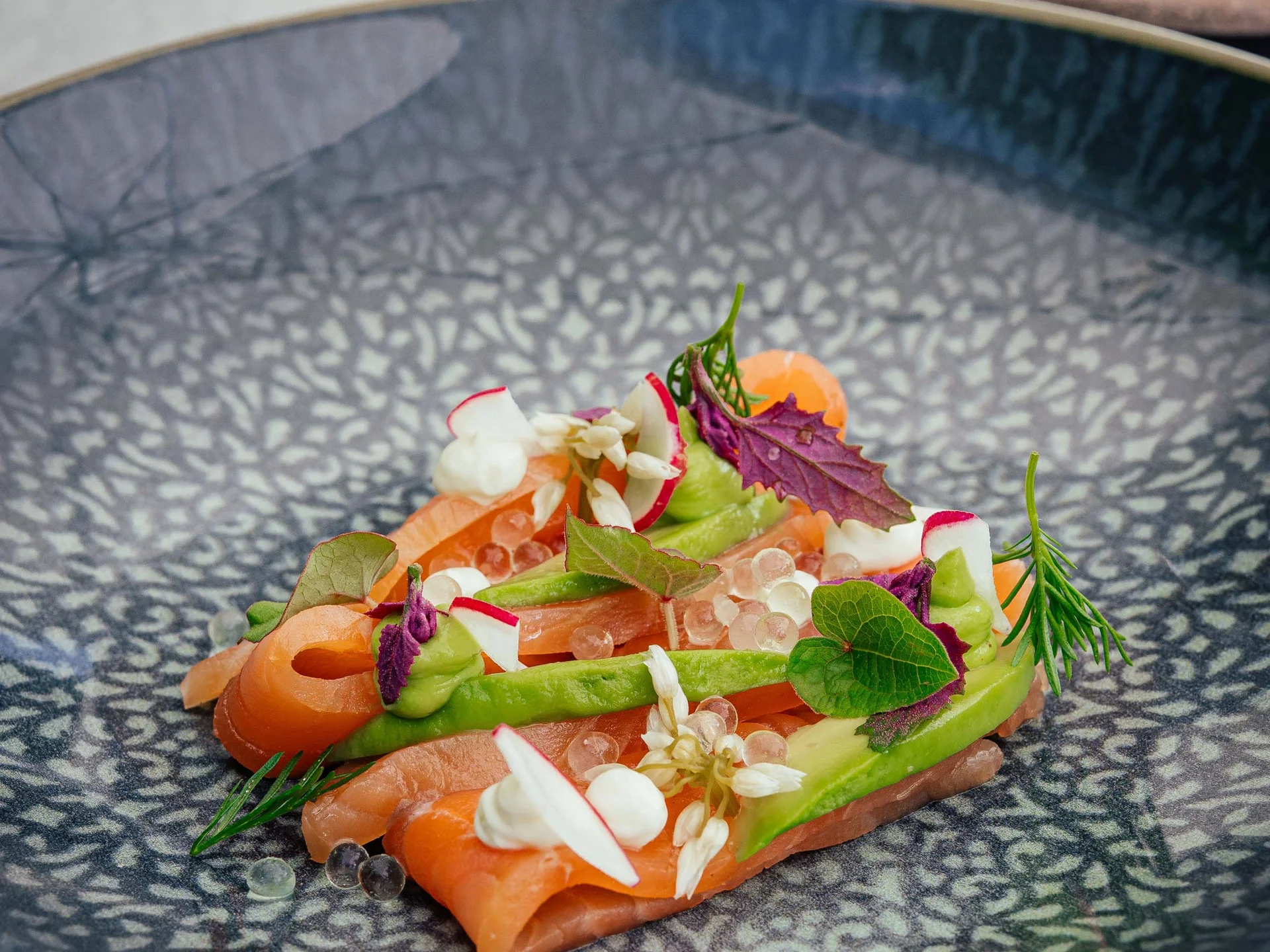 Der Lodenwirt in Vintl | Hotel, Residence & Restaurant Fein angerichteter Lachs mit Kräutern und essbaren Blüten auf blauem Teller